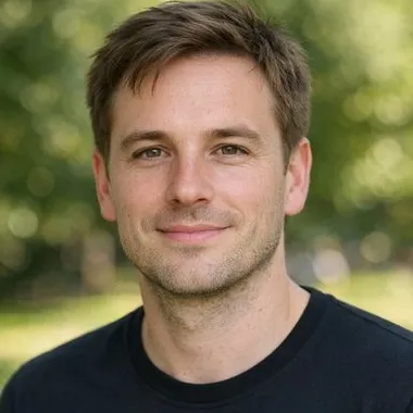 Young man, light brown hair, hazel eyes, dark crew-neck, sunlit park behind